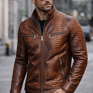 Puede incluir: Chaqueta de cuero marrón con textura de piel de serpiente, con cremallera frontal, múltiples bolsillos con cremallera y cuello alzado. La chaqueta se lleva con pantalones negros y gafas de sol.