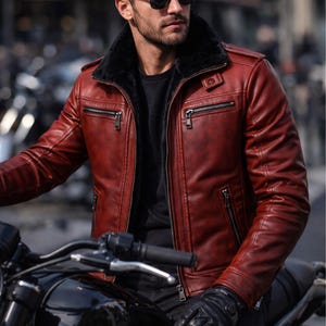 May include: A red leather jacket with a black fur collar and multiple zippered pockets. The jacket is worn with black gloves and sunglasses. The DRESSORT logo is visible in the upper left corner. The jacket is worn while sitting on a motorcycle.