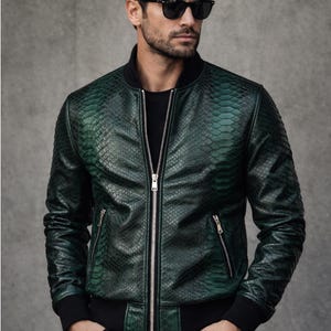 Może przedstawiać: Zielona kurtka bomberka o fakturze skóry węża z czarnym kołnierzem, mankietami i dołem. Kurtka posiada srebrny zamek błyskawiczny i dwie boczne kieszenie. Model nosi czarne okulary przeciwsłoneczne i czarne spodnie. Logo Dressort.