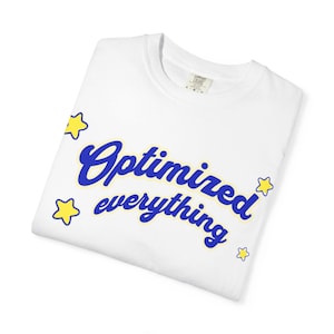 Peut inclure: T-shirt blanc plié avec l'inscription "Optimized everything" en bleu avec un contour jaune et des étoiles jaunes. Une étiquette est visible au niveau de l'encolure.
