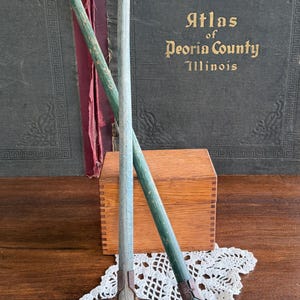 May include: Two antique miniature garden rakes with wooden handles painted blue and green, resting on a white lace doily. A wooden box and a vintage book titled "Atlas of Peoria County Illinois" are in the background.