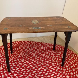 May include: A vintage wooden folding table with a painted top featuring a floral design. The table has dark brown legs and is sitting on a red and white braided rug.