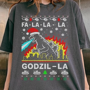 Può includere: T-shirt grigio scuro con un design festivo di Godzilla. La grafica include il testo "FA-LA-LA-LA" e "GODZIL-LA", un Godzilla con un cappello di Babbo Natale e uno skyline della città in fiamme. Il design presenta anche carri armati e fiocchi di neve.