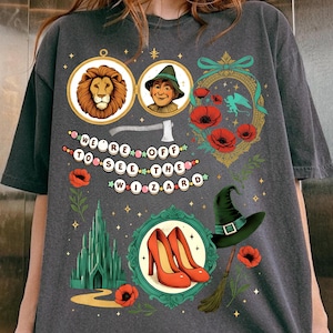 May include: Dark gray t-shirt featuring colorful illustrations from The Wizard of Oz. The design includes portraits of the Lion and Scarecrow, a green witch hat, red shoes, and the text "WIZARD". The shirt has a relaxed fit.