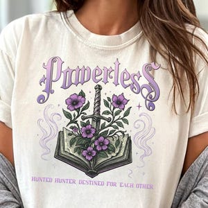 Może przedstawiać: Kremowa koszulka z motywem fantasy. Projekt przedstawia słowo "Powerless" w fioletowym skrypcie nad otwartą książką z mieczem i fioletowymi kwiatami. Tekst "Hunted Hunter Destined For Each Other" znajduje się poniżej.