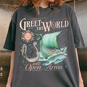 May include: Dark gray t-shirt featuring a vintage-style illustration of a Viking ship sailing on waves, with a sun and a harp. The text reads "Greet the World with Open Arms."