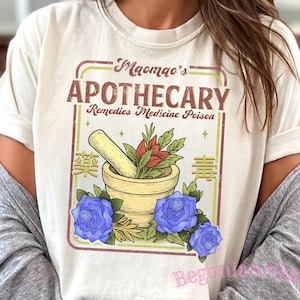 Puede incluir: Camiseta color crema con un diseño de boticario vintage. El gráfico incluye el texto "Maomao's Apothecary" con un mortero y una maja, elementos botánicos y rosas azules. También se incluyen las palabras "Remedies Medicine Poison".