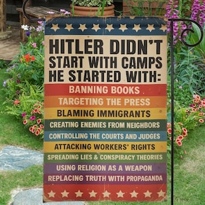 May include: A garden flag with the text "HITLER DIDN'T START WITH CAMPS HE STARTED WITH:" followed by a list of actions. The flag has a vintage design with a blue top, red and white stripes, and stars. It is displayed in a garden setting.