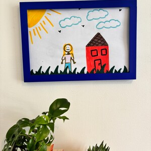 Puede incluir: Dibujo enmarcado con un borde azul. La obra de arte representa un sol amarillo, nubes azules y una casa roja con techo marr&oacute;n. Un personaje de palitos est&aacute; de pie cerca de la casa, y hierba verde en la parte inferior.