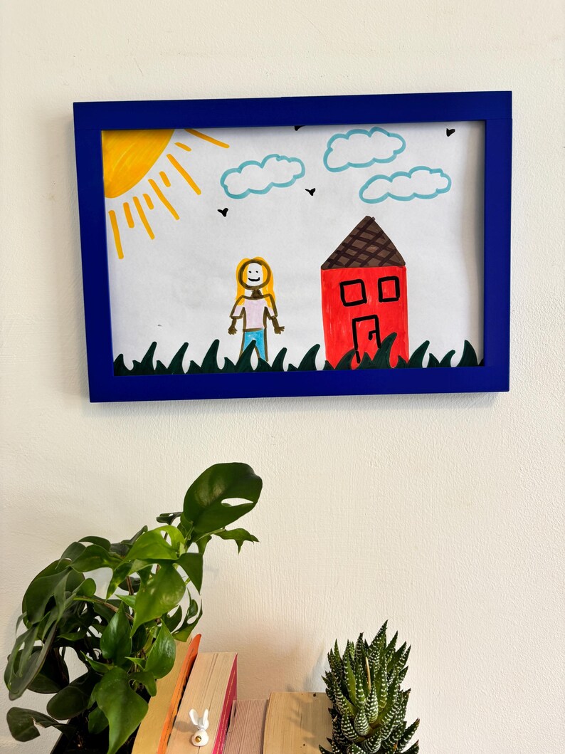 Puede incluir: Dibujo enmarcado con un borde azul. La obra de arte representa un sol amarillo, nubes azules y una casa roja con techo marr&oacute;n. Un personaje de palitos est&aacute; de pie cerca de la casa, y hierba verde en la parte inferior.