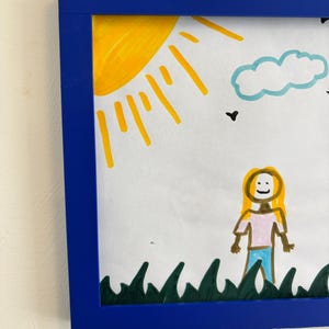 Puede incluir: Dibujo enmarcado con un borde azul. La obra de arte representa un sol amarillo, una nube azul y una persona con cabello rubio, una camisa rosa y pantalones azules. Hierba verde dibujada en la parte inferior de la imagen.