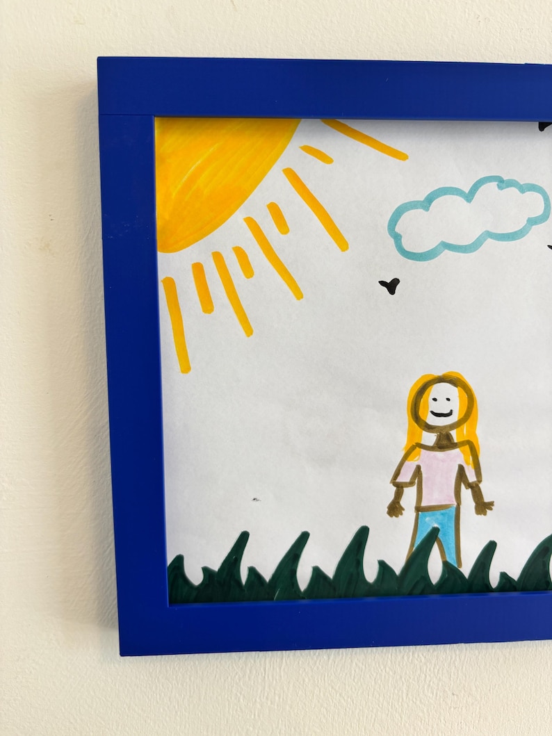 Puede incluir: Dibujo enmarcado con un borde azul. La obra de arte representa un sol amarillo, una nube azul y una persona con cabello rubio, una camisa rosa y pantalones azules. Hierba verde dibujada en la parte inferior de la imagen.