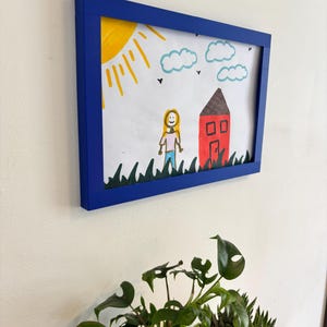 Puede incluir: Dibujo infantil enmarcado en un marco azul. Representa una casa roja con tejado marr&oacute;n, sol amarillo y nubes azules. Tambi&eacute;n se ve una persona y hierba verde.