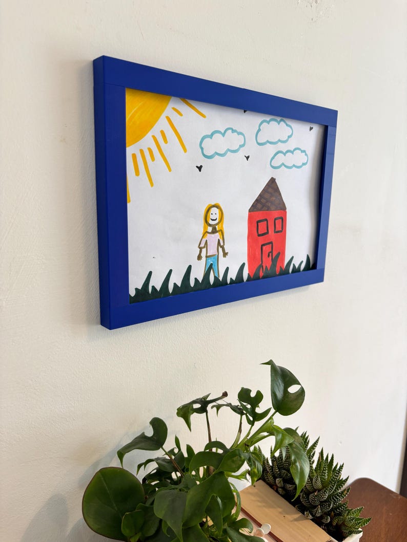 Puede incluir: Dibujo infantil enmarcado en un marco azul. Representa una casa roja con tejado marr&oacute;n, sol amarillo y nubes azules. Tambi&eacute;n se ve una persona y hierba verde.