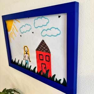 Puede incluir: Un dibujo enmarcado con un borde azul. La obra de arte representa una casa roja, un sol amarillo, nubes azules y una persona de pie en hierba verde. El dibujo est&aacute; sobre papel blanco.
