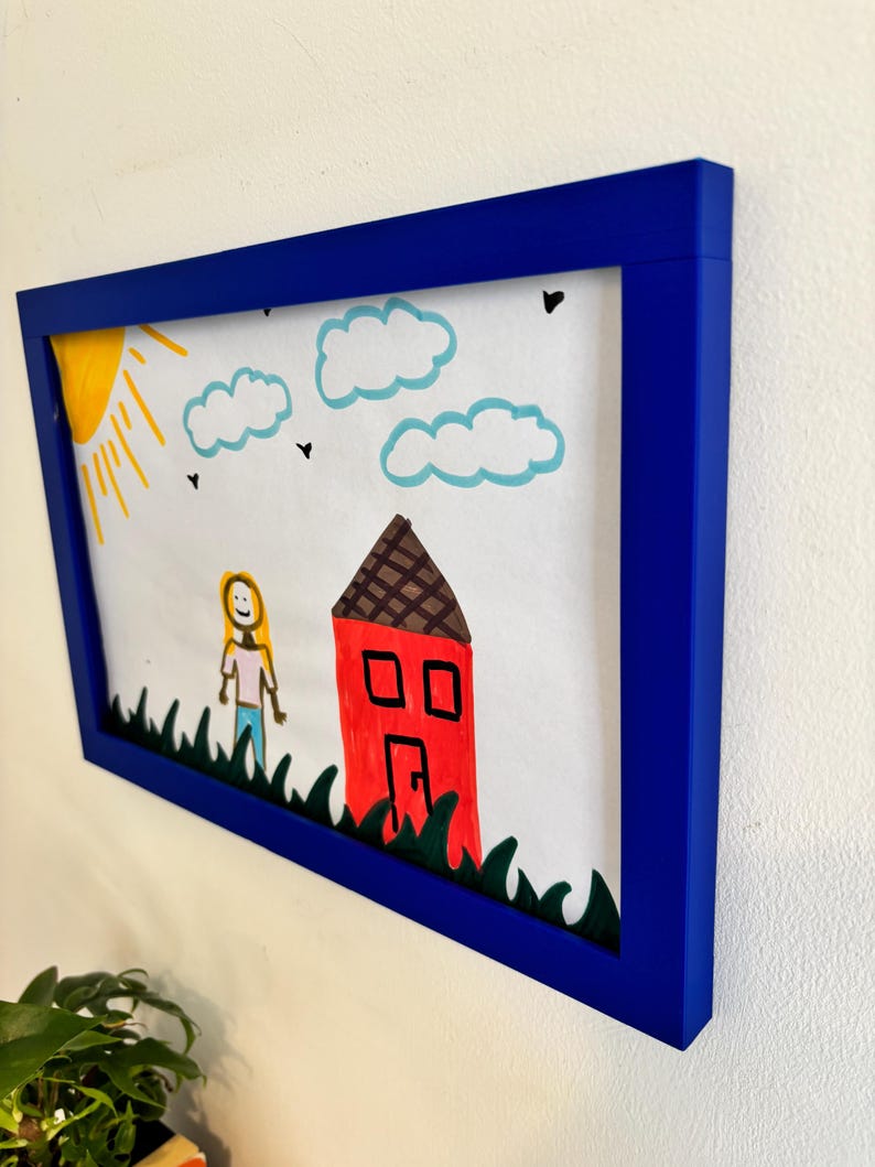 Puede incluir: Un dibujo enmarcado con un borde azul. La obra de arte representa una casa roja, un sol amarillo, nubes azules y una persona de pie en hierba verde. El dibujo est&aacute; sobre papel blanco.