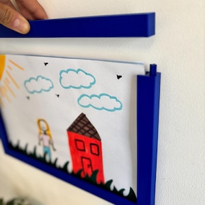 Peut inclure: Un cadre photo bleu contenant un dessin d'enfant représentant une maison, des nuages et une personne. Le cadre est présenté contre un mur blanc. Le dessin est aux couleurs vives.