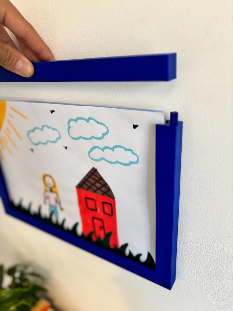 Puede incluir: Un marco de fotos azul que contiene un dibujo infantil de una casa, nubes y una persona. El marco se sostiene contra una pared blanca. El dibujo es de colores brillantes.