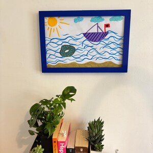 Puede incluir: Un dibujo infantil enmarcado de un barco navegando sobre agua azul ondulada bajo un sol amarillo y nubes azules. Un objeto verde flota en el agua. El marco es azul. Debajo hay libros y plantas.