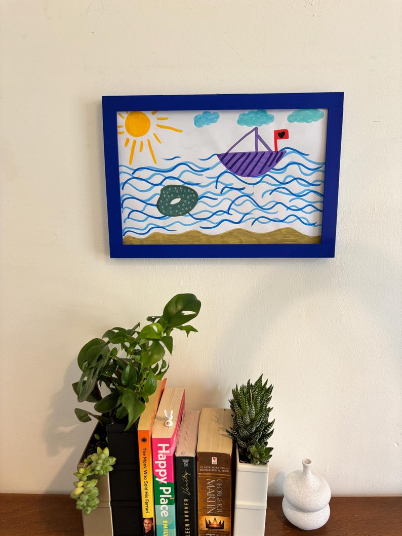 Puede incluir: Un dibujo infantil enmarcado de un barco navegando sobre agua azul ondulada bajo un sol amarillo y nubes azules. Un objeto verde flota en el agua. El marco es azul. Debajo hay libros y plantas.