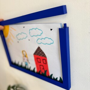 Puede incluir: Un marco de fotos azul que contiene un dibujo infantil de una casa, una persona, un sol y nubes. El dibujo est&aacute; hecho con crayones y rotuladores. El marco es de color azul oscuro y est&aacute; montado en una pared blanca.
