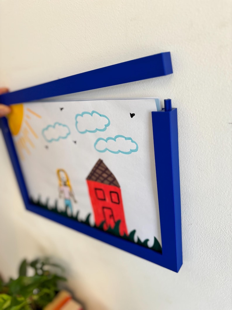 Puede incluir: Un marco de fotos azul que contiene un dibujo infantil de una casa, una persona, un sol y nubes. El dibujo est&aacute; hecho con crayones y rotuladores. El marco es de color azul oscuro y est&aacute; montado en una pared blanca.