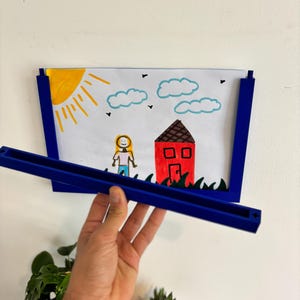 Puede incluir: Un marco de fotos azul que contiene un dibujo infantil de una casa, un sol, nubes y una persona. El marco est&aacute; sostenido por una mano. El dibujo presenta una casa roja, un sol amarillo y un cielo azul.