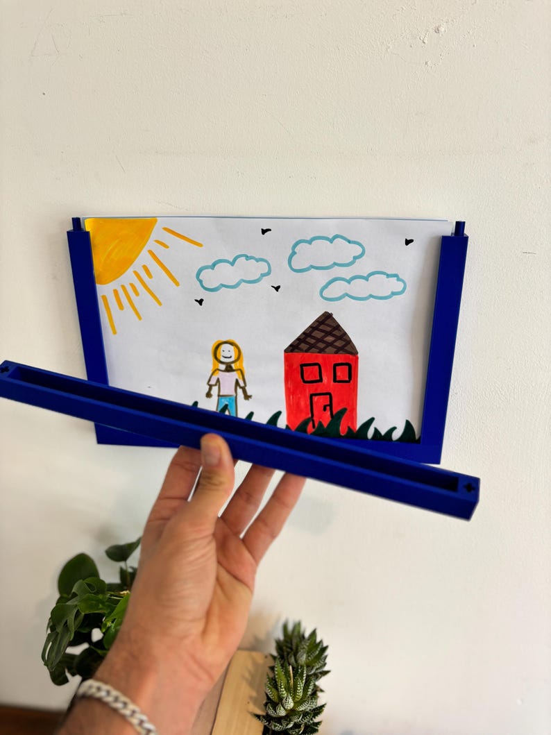 Puede incluir: Un marco de fotos azul que contiene un dibujo infantil de una casa, un sol, nubes y una persona. El marco est&aacute; sostenido por una mano. El dibujo presenta una casa roja, un sol amarillo y un cielo azul.