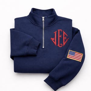 Könnte beinhalten: Marineblaues Sweatshirt mit Viertelreißverschluss, rotem Monogramm und einem amerikanischen Flaggenaufnäher auf dem Ärmel. Das Sweatshirt hat einen gerippten Kragen und Bündchen sowie einen silbernen Reißverschluss.
