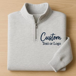 May include: A pale grey quarter-zip sweatshirt, folded neatly. It features the word "Custom" and the text "Text or Logo" in navy blue. The design highlights the zipper and ribbed cuffs.