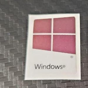 Puede incluir: Un cuadrado blanco con la palabra "Windows®" impresa. El cuadrado presenta un diseño de cuatro paneles en tonos de rosa y rojo. El fondo es una superficie texturizada de color gris oscuro.