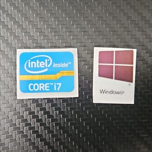 Puede incluir: Dos pegatinas pequeñas sobre una superficie negra texturizada. Una pegatina tiene el logotipo de Intel Inside y el texto "CORE i7" en azul y amarillo. La otra pegatina tiene el logotipo de Windows y un diseño cuadrado granate.