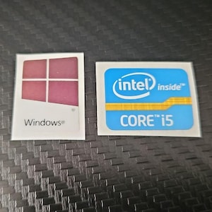 Puede incluir: Dos pegatinas de componentes informáticos. Una es un cuadrado blanco con un logotipo de Windows rojo y rosa. La otra es un cuadrado azul con el texto "intel inside" y "CORE i5".