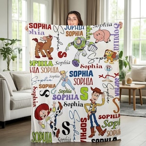 May include: White blanket featuring various Toy Story characters and the name "Sophia" in multiple fonts and colors. Characters include Woody, Buzz Lightyear, Jessie, and others. The blanket is held up by a person.