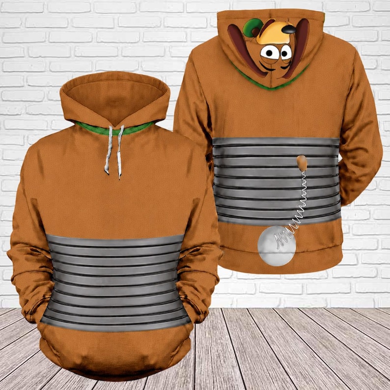 May include: A brown hoodie featuring a graphic print of a cartoon dog character. The design includes a grey horizontal ribbed pattern across the midsection, a hood with dog ears, and a small white ball attached by a spring on the back.