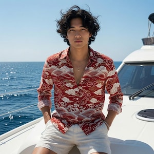 May include: A long-sleeved button-down shirt in a deep red hue featuring a repeating white and gold pattern of stylized clouds and ocean waves. The shirt is paired with light beige shorts and layered metallic necklaces with circular pendants.
