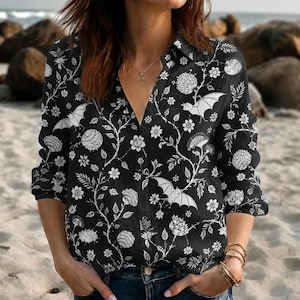 May include: A long-sleeved button-down shirt featuring a black base with a white botanical print of flowers, vines, and flying bats. The shirt has a collared neckline and a front button placket, showcasing a repeating pattern of white floral elements.