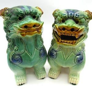 May include: Two ceramic Foo Dog statues. The statues are green with blue and brown accents. They have detailed features, including open mouths, and are posed in a seated position. The Foo Dogs are a traditional Asian decorative item.