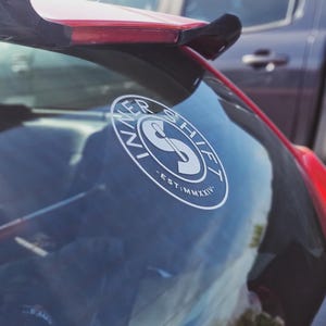 Peut inclure: Autocollant circulaire blanc avec les mots "INNER SHIFT" sur le bord et un "S" stylisé au centre. L'autocollant est sur une vitre de voiture bleu foncé, avec le texte "EST:MMXXIV" en bas.