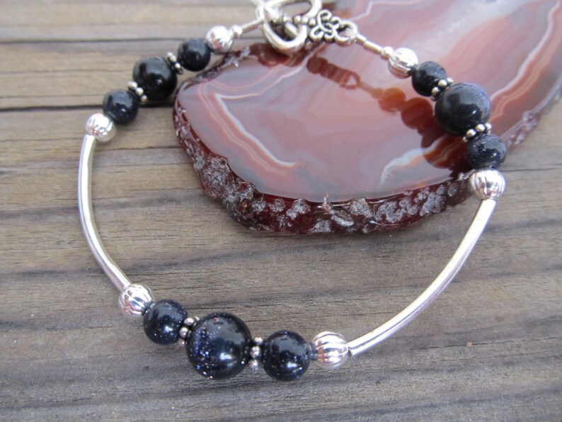 May include: A silver bracelet featuring dark blue and black beads. The bracelet has a curved silver bar design with a toggle clasp. The beads have a speckled appearance, and the bracelet is displayed on a wooden surface.