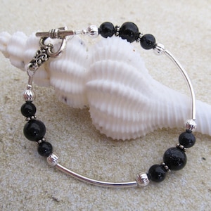 May include: A silver bracelet with black beads and a toggle clasp. The bracelet features a curved silver bar and is adorned with various sizes of black beads. The clasp has an ornate design. The bracelet is displayed on a white seashell.