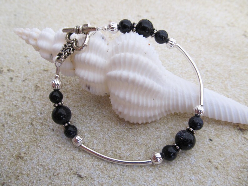 May include: A silver bracelet with black beads and a toggle clasp. The bracelet features a curved silver bar and is adorned with various sizes of black beads. The clasp has an ornate design. The bracelet is displayed on a white seashell.