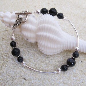 May include: A silver bracelet with black beads and a toggle clasp. The bracelet features a curved design with alternating black beads and silver accents. The toggle clasp has an ornate design. The bracelet is displayed on a white seashell.