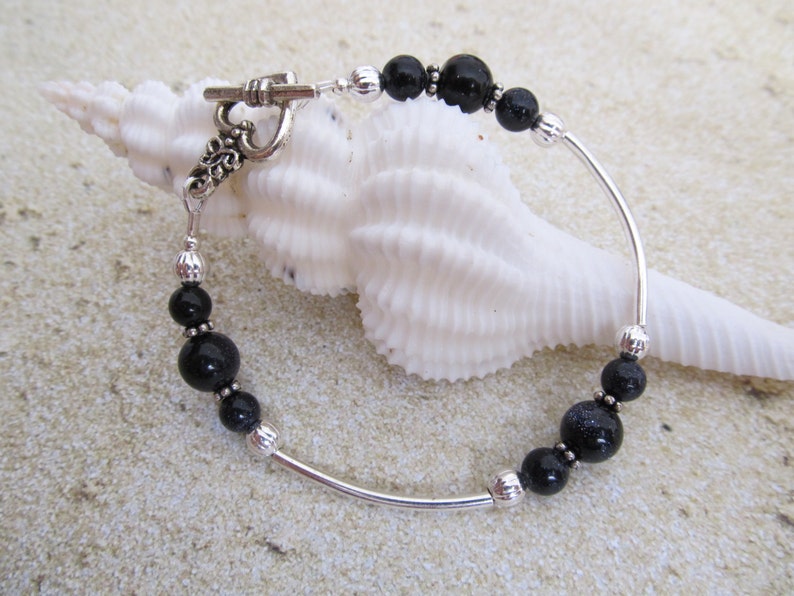 May include: A silver bracelet with black beads and a toggle clasp. The bracelet features a curved design with alternating black beads and silver accents. The toggle clasp has an ornate design. The bracelet is displayed on a white seashell.
