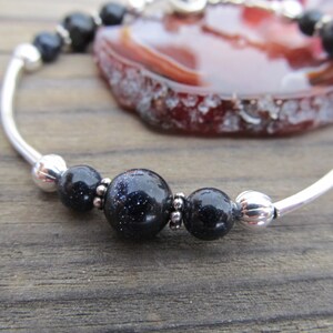 May include: A silver bracelet featuring dark blue, speckled beads and smaller black beads. The bracelet has a curved, open design with a polished finish. The background includes a slice of a red and brown stone.
