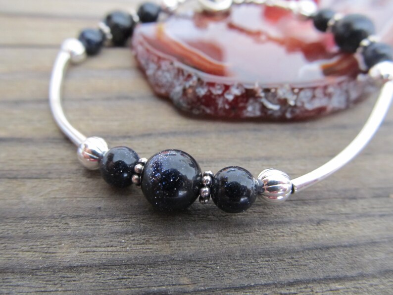 May include: A silver bracelet featuring dark blue, speckled beads and smaller black beads. The bracelet has a curved, open design with a polished finish. The background includes a slice of a red and brown stone.