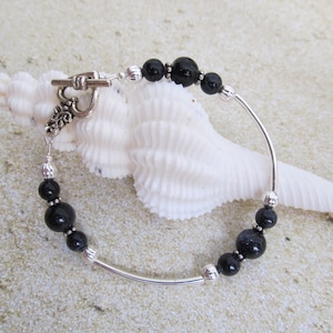 May include: A beaded bracelet featuring black and silver beads strung on a curved silver bar. The bracelet has a toggle clasp and is displayed on a white seashell against a sandy background. The beads vary in size and texture.