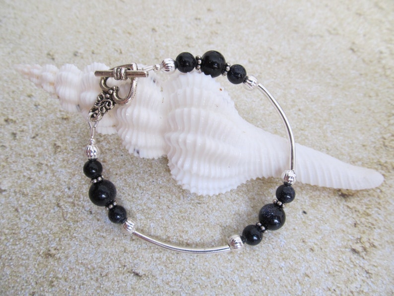 May include: A beaded bracelet featuring black and silver beads strung on a curved silver bar. The bracelet has a toggle clasp and is displayed on a white seashell against a sandy background. The beads vary in size and texture.