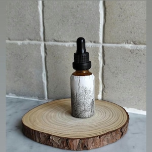 Puede incluir: Pequeña botella de vidrio ámbar con cuentagotas negro, envuelta con una etiqueta blanca con un diseño de árboles negros y grises. Descansa sobre una rodaja de madera redonda.
