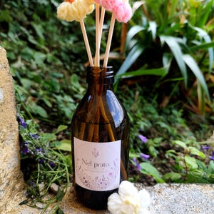 Può includere: Una bottiglia di vetro marrone con un'etichetta bianca "Nel prato" contiene bastoncini di legno sormontati da fiori di filato giallo, rosa e marrone. Un fiore bianco all'uncinetto è davanti alla bottiglia, con uno sfondo verde.